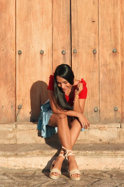 Genç bir bayan, yaz günü kırmızı bir gömlek, açık mavi bir etek ve bej renkli sandaletler giyerek büyük bir klasik kapının yanında oturuyor.