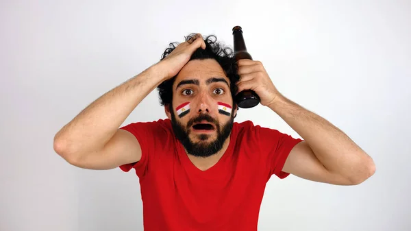 Shot of fan is disappointed with the team of his country. Man with the flag of Egypt makeup on his face and red t-shirt.