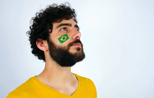 Sport fan head high and feeling proud when listening to the anthem of his country. Man with the flag of Brazil makeup on his face and yellow t-shirt.