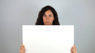 Beautiful woman with a serious expression on her face holding a blank white sign board in her hands in front of her chest with copy space for your text or advertising, over a grey background clipart