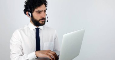 Çağrı merkezi gri arka plan üstünde laptop ile kulaklıklar, yakışıklı danışman. Genç ve esmer adam laptop ciddi bakan.