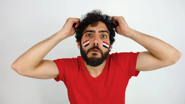 Sport fan disappointed with the team of his country. Man with the flag of Egypt makeup on his face and red t-shirt.