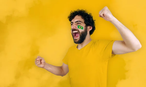 Sport fan screaming for the triumph of his team. Man with the flag of Brazil makeup on his face, yellow t-shirt and yellow background.