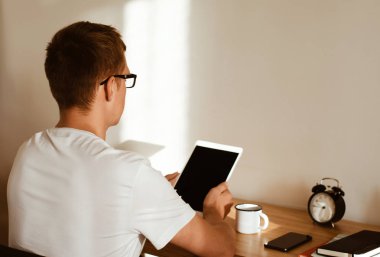 Masasında oturan genç bir adam, tablette kitap okuyor..