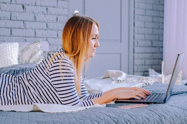 Beautiful girl in a striped blouse works at home on the bed behind a laptop.