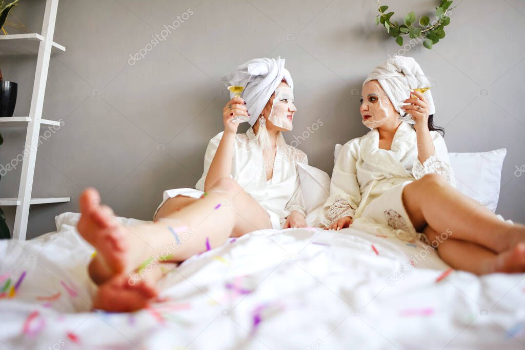Girlfriends with masks on their faces drink champagne while lying in bed. 