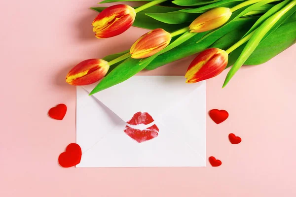 Valentines day background. Love letter concept. White envelope with red lipstick kiss, red hearts and bouquet of tulips on pink pastel.