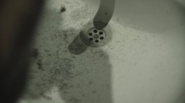 Adam banyo aynasının önünde tıraş makinesiyle sakalını tıraş ediyor. Saçlar lavaboya dökülüyor.