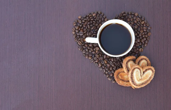Heart of coffee beans with a white mug in it and confectionery on a dark background