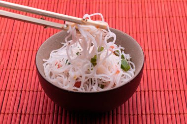 Pirinç erişte ve sebze ile Asya salatası. Kırmızı ahşap arka plan üzerinde izole çubukları ile siyah ve kahverengi bir kapta Pişmiş pirinç şehriye Tay tarzı. 