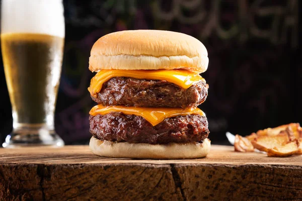 Rustik patates kızartması ve serin bir bardak bira ile ahşap rustik masada taze çift burger closeup. Lezzetli çizburger. Ev yapımı hamburger, biftek ve peynir. Koyu arka plan