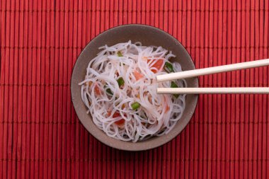 Pirinç erişte ve sebze ile Asya salatası. Kırmızı ahşap arka plan üzerinde izole çubukları ile siyah ve kahverengi bir kapta Pişmiş pirinç şehriye Tay tarzı. 