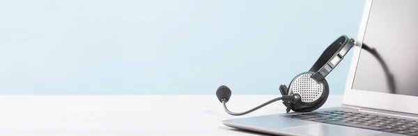 Masa mavi arka planında kulaklık olan boş ekranlı bir dizüstü bilgisayar parçası. kopyalama alanı. Uzaktan öğrenme ya da evden çalışma, online kurslar. Yardım masası veya çağrı merkezi minima konsept pankartı