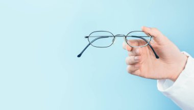 Kadın doktor göz doktoru veya göz doktoru ince metal çerçeveli bir gözlük tutuyor. Minimum afiş, mavi arka plan. Uzayı kopyala Optik ya da göz doktoru, göz testi ve kontroller.