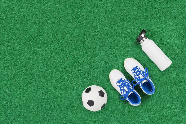 Soccer ball, cleats and water bottle on green artificial turf, top view with copy space, football concept. Game or sports club class and tournament concept