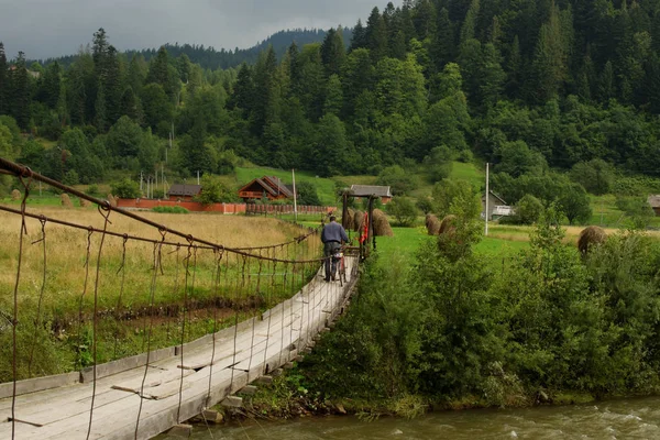 Man with bicycle crossing old suspension bridge over the mountain river. Beautiful rustic landscape view on Carpathian hills. Ukrainian village. Green tourism concept.