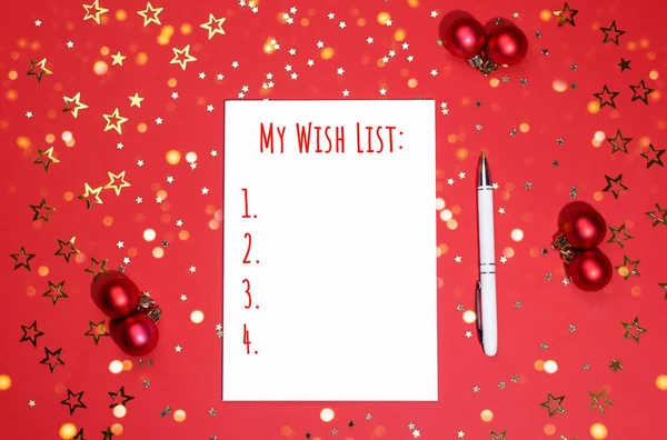 Notebook with pen for writing a wish list on vibrant red background decorated with Christmas baubles, star-shaped confetti and festive lights. New Year and Christmas wishing concept.