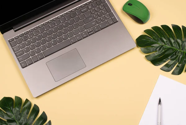 Beautiful shiny artificial monstera leaves, note pad with white pen for writing, green wireless mouse and laptop on pale yellow background. Copy space, flat lay style. Home office workspace.