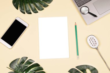 Güzel parlak yapay monstera yaprakları, Not defteri ile yazma, beyaz smartphone, modern Led lamba, büyüteç ve uçuk sarı arka plana laptop için yeşil kalem.