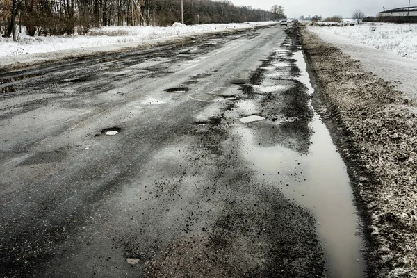 road is paved with cracks in pits and puddles from the snow. Landscape of the winter road in cloudy gray weather. Concept of road repair, opacity for the driver of the car