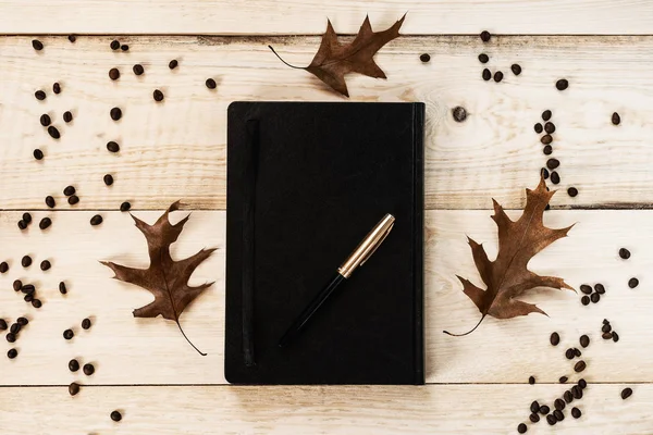 Black notebook with a pen, three fallen leaves and coffee beans on a wooden board.
