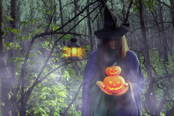 Young witch in the dark forest on Halloween. Girl dressed as a witch with Jack-o-lantern in a misty forest at night.