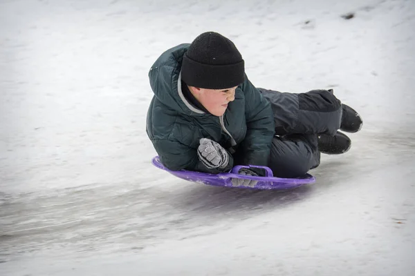 In winter, in a forest, a boy lying on his stomach slides down a — Stock Photo, Image