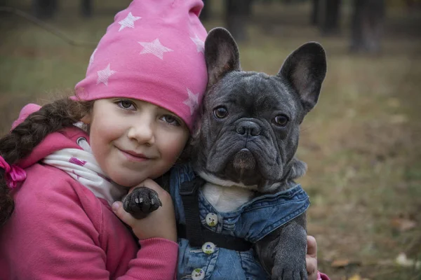 In the autumn forest, a little girl holds a French bulldog in he — Stock Photo, Image