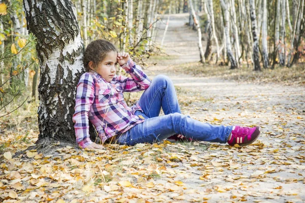 In the autumn birch forest, a little sad upset girl sits under a — Stock Photo, Image