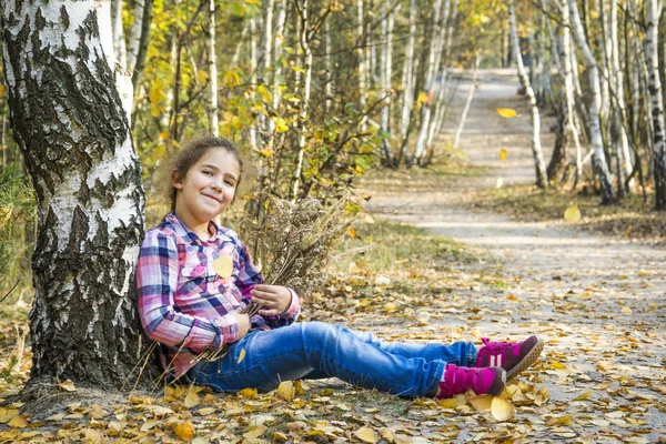 V podzimním břízovém lese sedí malá holčička pod stromem poblíž — Stock fotografie