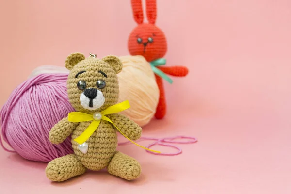 A brown knitted bear sits on a pink background, behind it is a ball of pink and yellow yarn, and an orange knitted hare stands behind the yarn. There is a place for text.