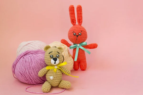 Ein brauner Strickbär sitzt auf einem rosafarbenen Hintergrund, dahinter ein Knäuel aus rosa und gelben Garnen, und hinter dem Garn steht ein orangefarbener gestrickter Hase. Es gibt einen Platz für Text. — Stockfoto