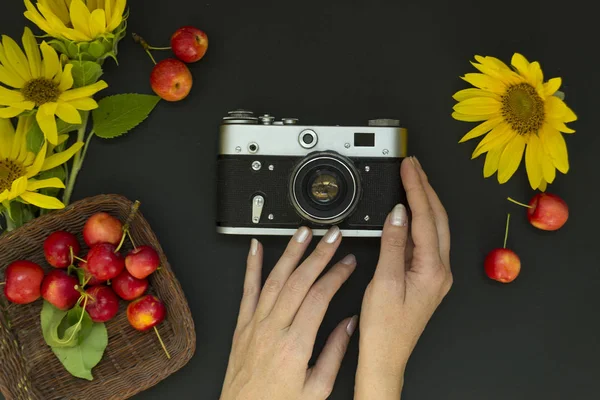 Weibliche Hände mit einer Filmkamera auf schwarzem Hintergrund. In der Nähe befinden sich gelbe Sonnenblumenblüten und reife rote Äpfel. Konzeptsommer. — Stockfoto