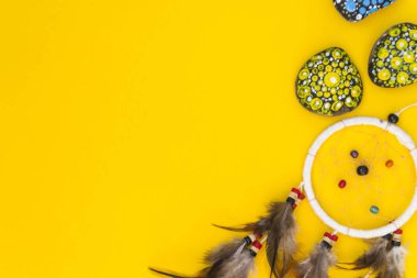 Kahverengi kuş tüylü dreamcatcher, ve iplikleri ve ip asılı boncuklar ile. Dreamcatcher el yapımı. Boyalı renkli taşlar arasında sarı bir arka plan üzerinde yatıyor.