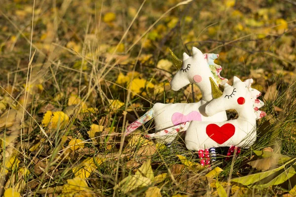 Two textile toys unicorns stand on the yellow foliage and grass, graze. Close-up. Blurred background.