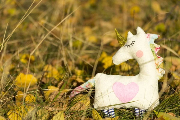 One textile toy unicorn stands on yellow foliage and grass, graze. Close-up. Blurred background. There is a place for text.