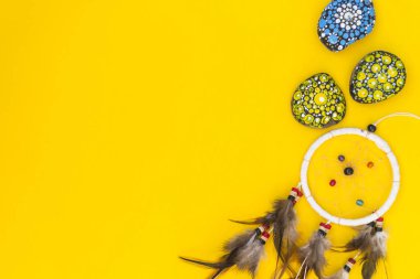 Kahverengi kuş tüylü dreamcatcher, ve iplikleri ve ip asılı boncuklar ile. Dreamcatcher el yapımı. Boyalı renkli taşlar arasında sarı bir arka plan üzerinde yatıyor.