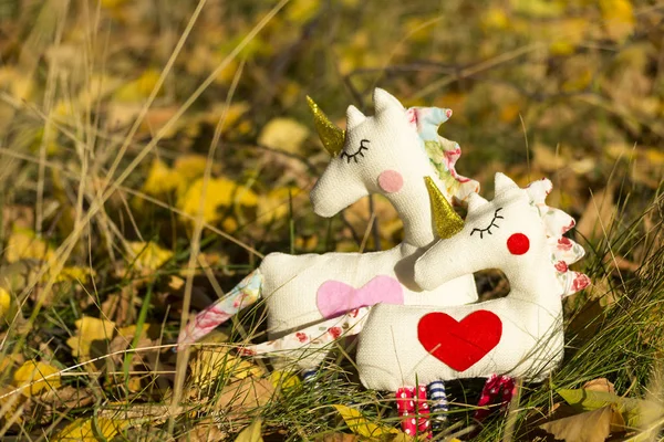 Two textile toys unicorns stand on the yellow foliage and grass, graze. Close-up. Blurred background.