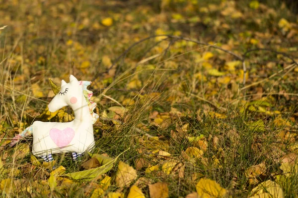 One textile toy unicorn stands on yellow foliage and grass, graze. Close-up. Blurred background. There is a place for text.