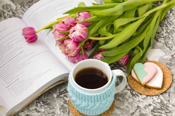 White cup of tea in a knitted blue case with an open book and with pink flowers tulips and sweets, with gingerbread on a textural table.