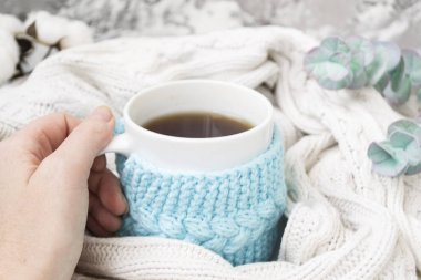 Bir kadın el örme mavi çerçeve içinde çay beyaz bir kupa tutuyor ve beyaz örme ekose sarılmış. Arka planda, okaliptüs ve pamuk çipler. Yatakta kahvaltı. Rahat.