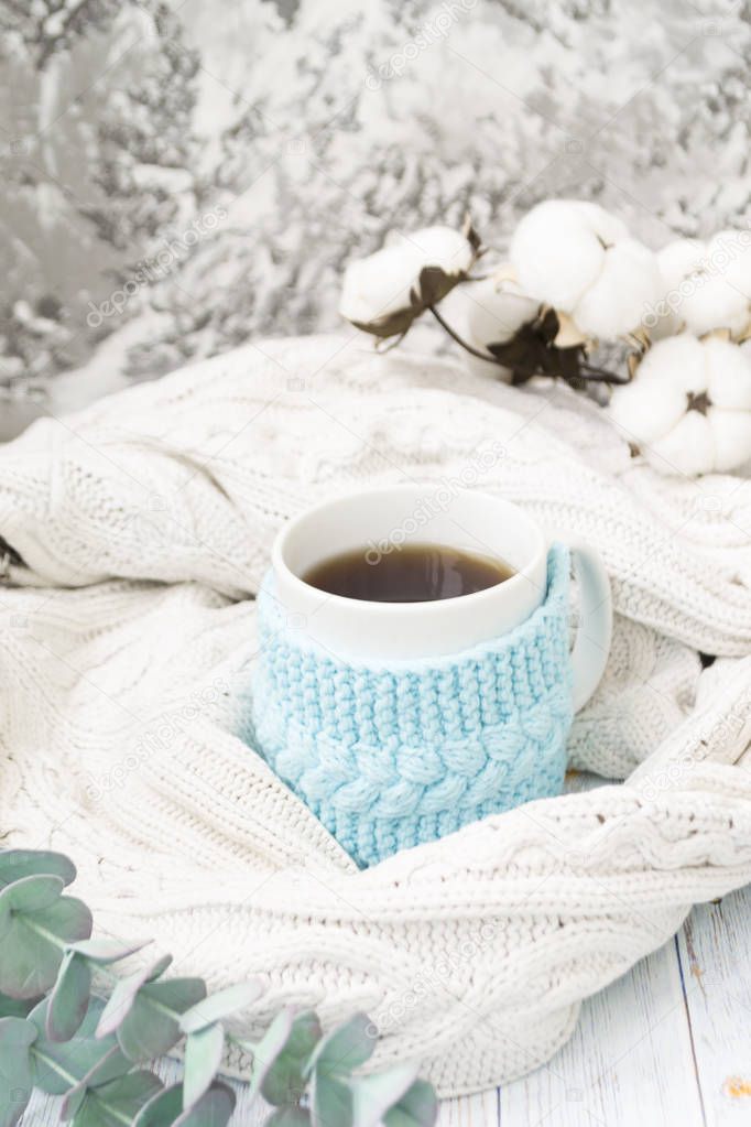 White mug with tea in a knitted blue frame and wrapped in a white knitted plaid. In the background, sprigs of eucalyptus and cotton. Breakfast in bed. Cozy.