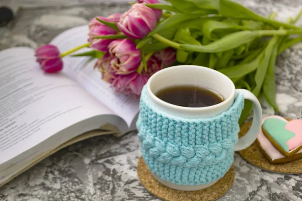 Açık bir kitap ile bir örme mavi durumda çay beyaz fincan ve pembe çiçekler ile lale ve tatlılar, bir dokusal masada Gingerbread ile.