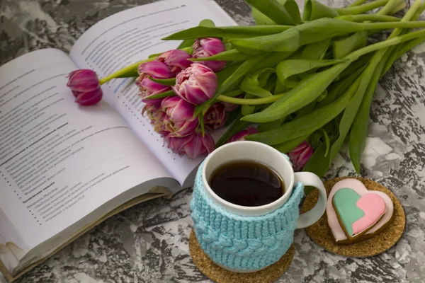 Taza blanca de té en una caja azul de punto con un libro abierto y con flores rosas tulipanes y dulces, con pan de jengibre en una mesa textural . —  Fotos de Stock