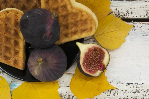 Плоды инжира на тарелке лежат с печеньем в форме сердца в тарелке. Вокруг плиты жёлтые листья деревьев. Фон белый, деревянный. Осенняя концепция . — стоковое фото