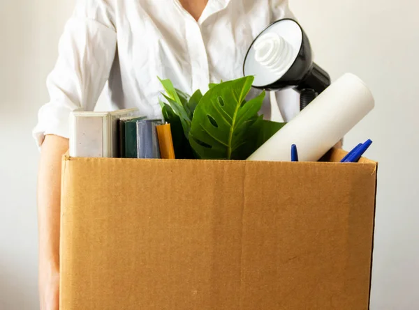 lose job concept. Hand holds box of fired worker. In a box they lay documents, folders, a flower, stationery, a table lamp. Woman worker in a white shirt holds a box.