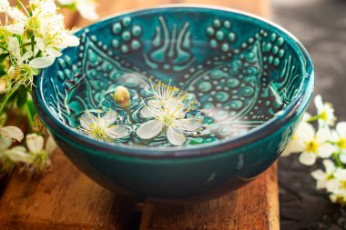 Kahverengi ahşap bir tahtanın üzerinde su dolu mavi bir fincan duruyor. Yakınlarda güzel beyaz kiraz çiçekleri tomurcukları var. Sakura 'nın güzel bahar kompozisyonu.