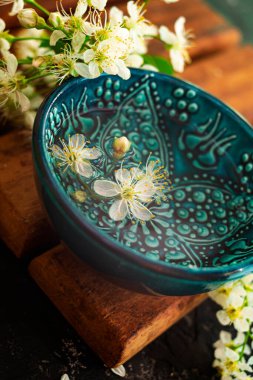 Kahverengi ahşap bir tahtanın üzerinde su dolu mavi bir fincan duruyor. Yakınlarda güzel beyaz kiraz çiçekleri tomurcukları var. Sakura 'nın güzel bahar kompozisyonu.