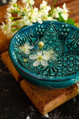 Kahverengi ahşap bir tahtanın üzerinde su dolu mavi bir fincan duruyor. Yakınlarda güzel beyaz kiraz çiçekleri tomurcukları var. Sakura 'nın güzel bahar kompozisyonu.