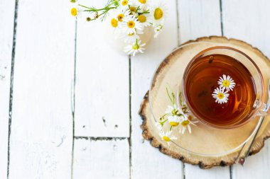Bir fincan papatya çayı ve beyaz tahta üzerinde papatya çiçekleri olan bitki çayı. Yakınlarda bir vazo beyaz çiçek duruyor. Boşluğu kopyala